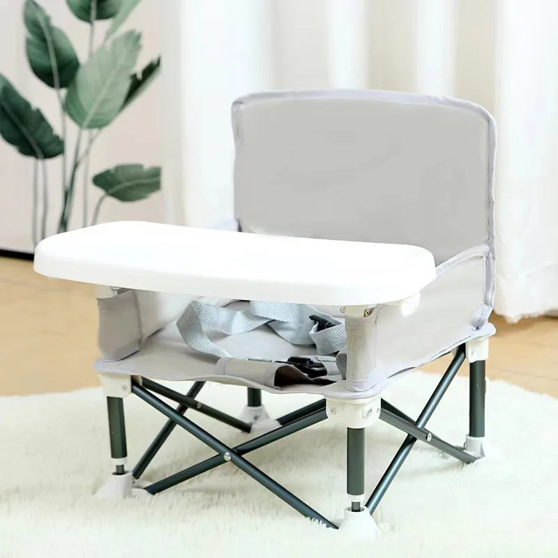 Foldable baby seat with tray on a white surface, plant and curtain in the background