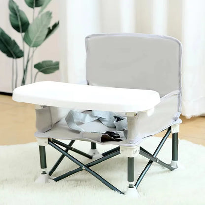 Foldable baby seat with tray on a white surface, plant and curtain in the background